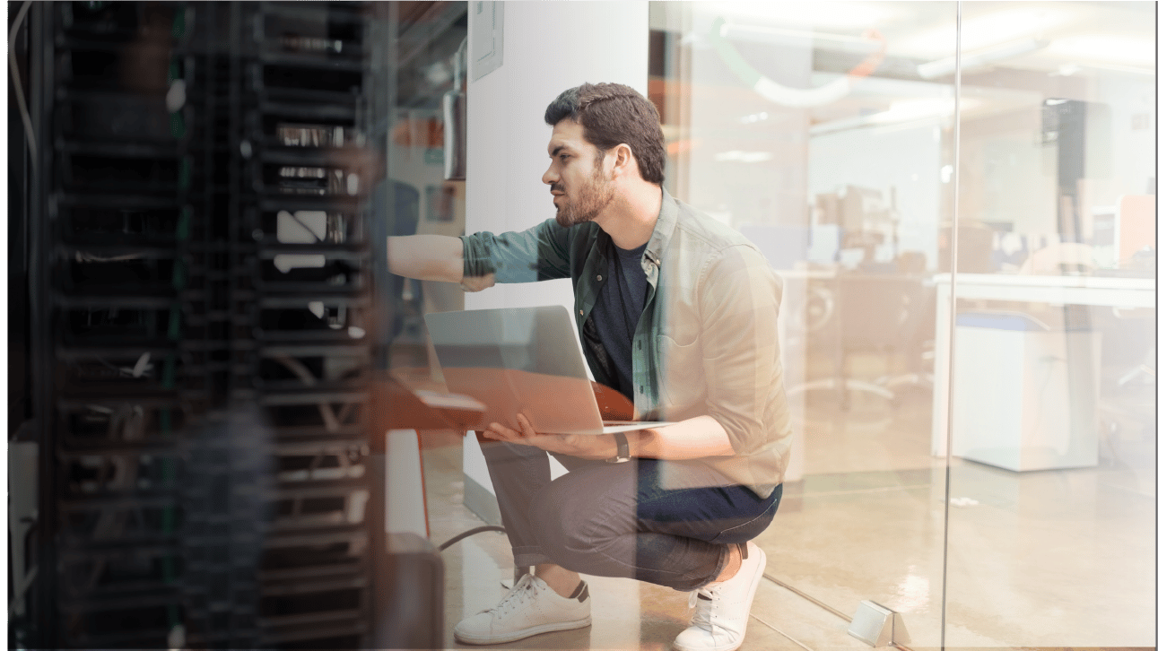 IT man kneels down to work on a data center rack while holding laptop.